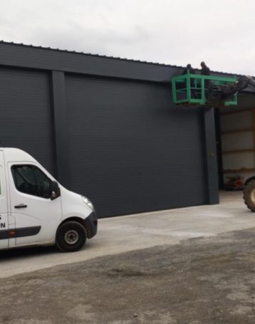 Chantier d’installation de portes sectionnelles sur hangar industriel gris avec engin de levage.
